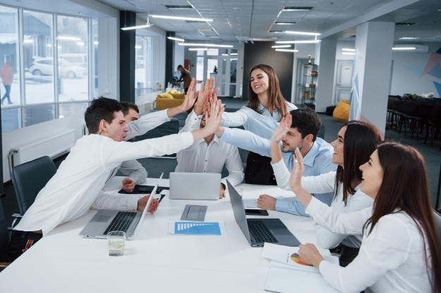Um grupo de pessoas no trabalho, em uma sala de reuniões, batendo as mãos um dos outros de forma a comemorar bons resultados. A motivação é um dos pilares da comunicação corporativa