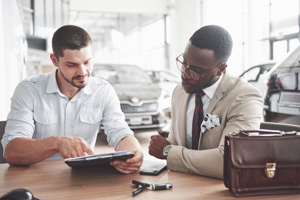 Imagem de dois homens sentados à mesa, um deles vende um carro e o outro compra